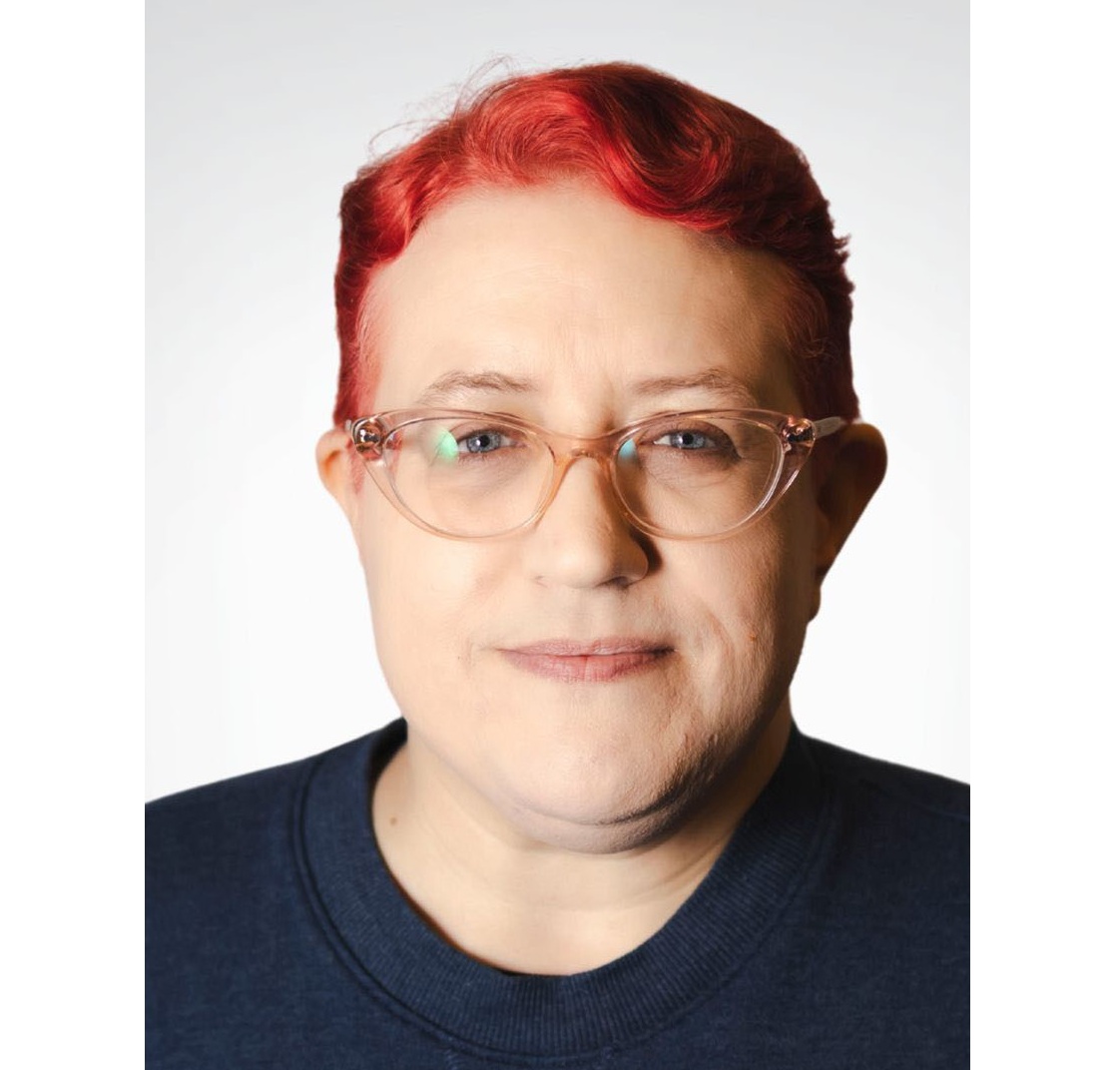 Photo of B.C. Skipalis, a fair-skinned person with clear-pink glasses, blue eyes, short, wavy dark coral-pink hair, and a navy blue sweater.